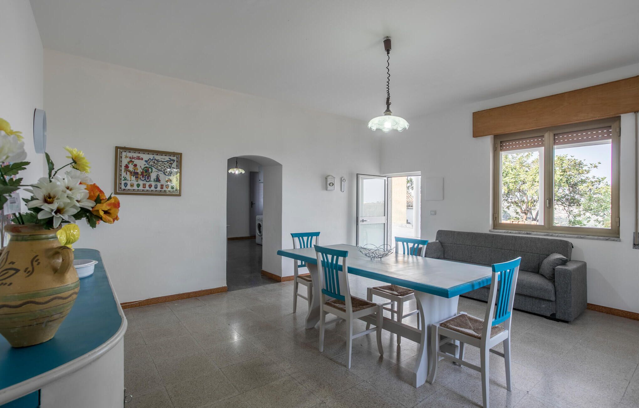 A dining area with a table and chairs, a sofa, a window with a view, and a hanging light fixture.