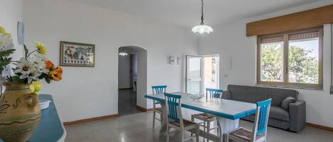 A dining area with a table and chairs, a sofa, a window with a view, and a hanging light fixture.