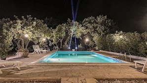 Une zone de piscine la nuit, avec un sol en pierre, des chaises longues et des arbres.