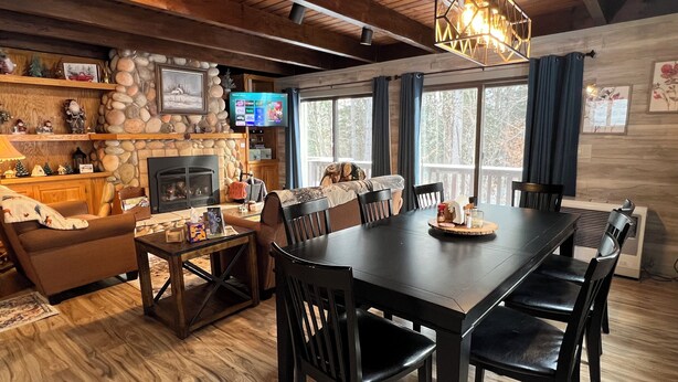 Un salon chaleureux avec une cheminée, un mur en pierre, un plafond en bois et une grande table à manger entourée de chaises.