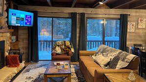 Un salon chaleureux avec une cheminée, un téléviseur diffusant des services de streaming et une vue sur une terrasse enneigée.