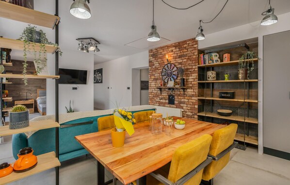 Un salon moderne avec une table à manger en bois, des chaises jaunes, un mur d’accent en briques et une bibliothèque remplie de livres et d’objets décoratifs.