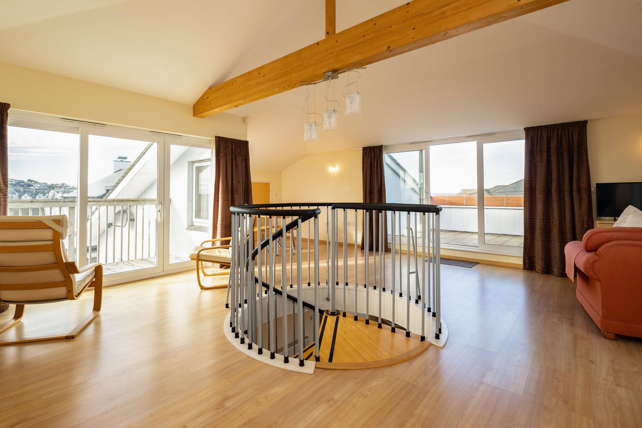 Un vaste salon doté d’un parquet, d’un escalier en colimaçon, d’une grande fenêtre offrant une vue dégagée et d’un canapé rouge.