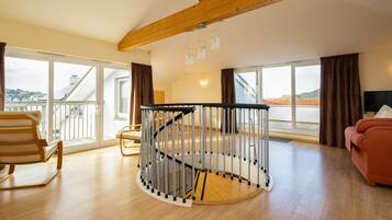 Un vaste salon doté d’un parquet, d’un escalier en colimaçon, d’une grande fenêtre offrant une vue dégagée et d’un canapé rouge.