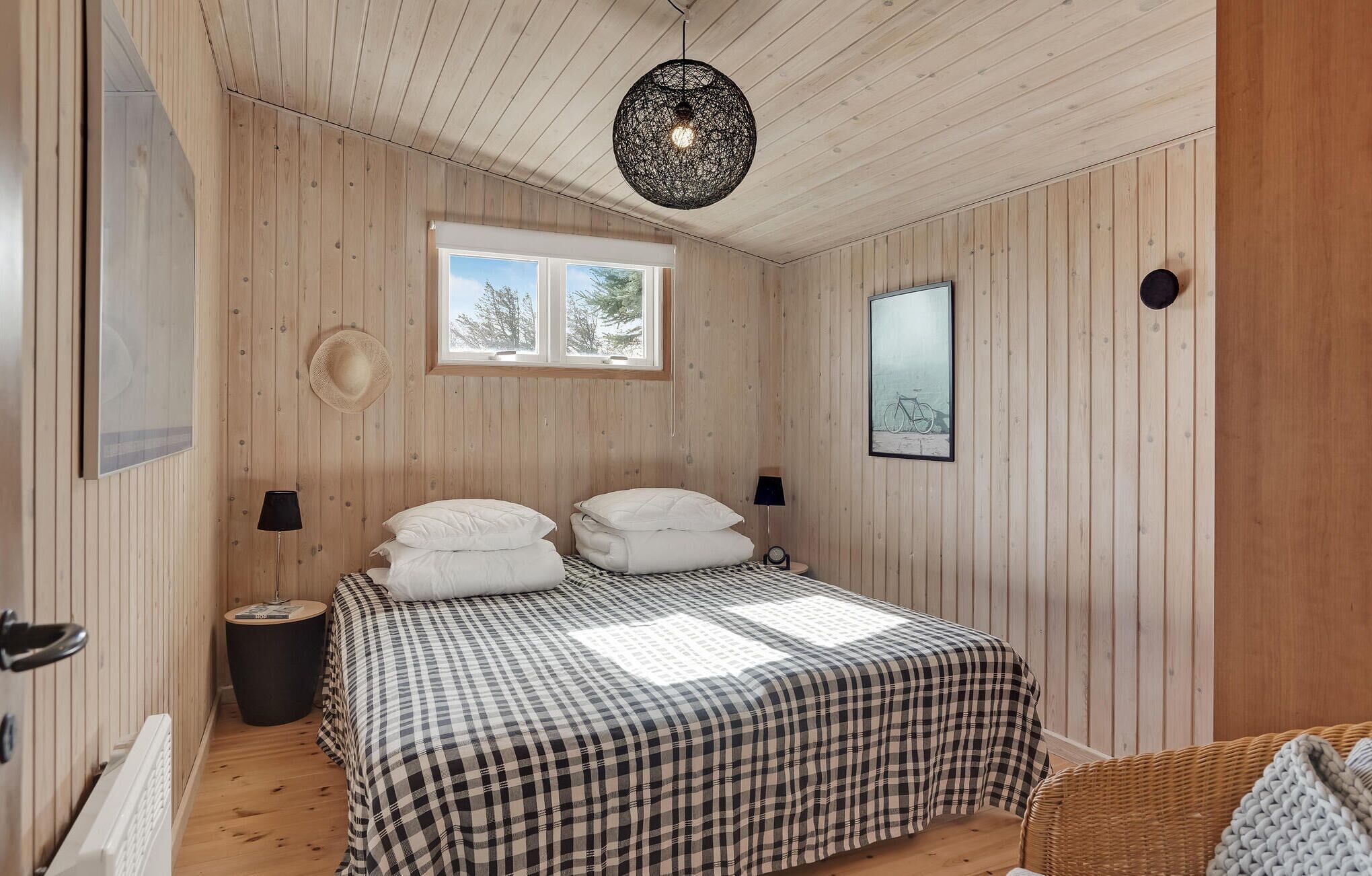 Una camera da letto con una coperta a quadretti, una finestra vista alberi, un cappello appeso al muro e una lampada rotonda.