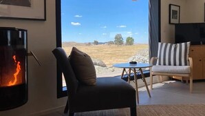A living room with a fireplace, a sofa, a striped chair, and a view of a dry landscape through a large window. - Farm stay accomodation (Pyalong)
