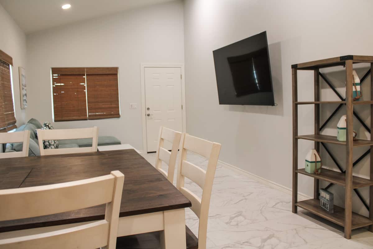 A dining area with a dark wooden table and white chairs, a television mounted on the wall, and a bookshelf with decorative items.