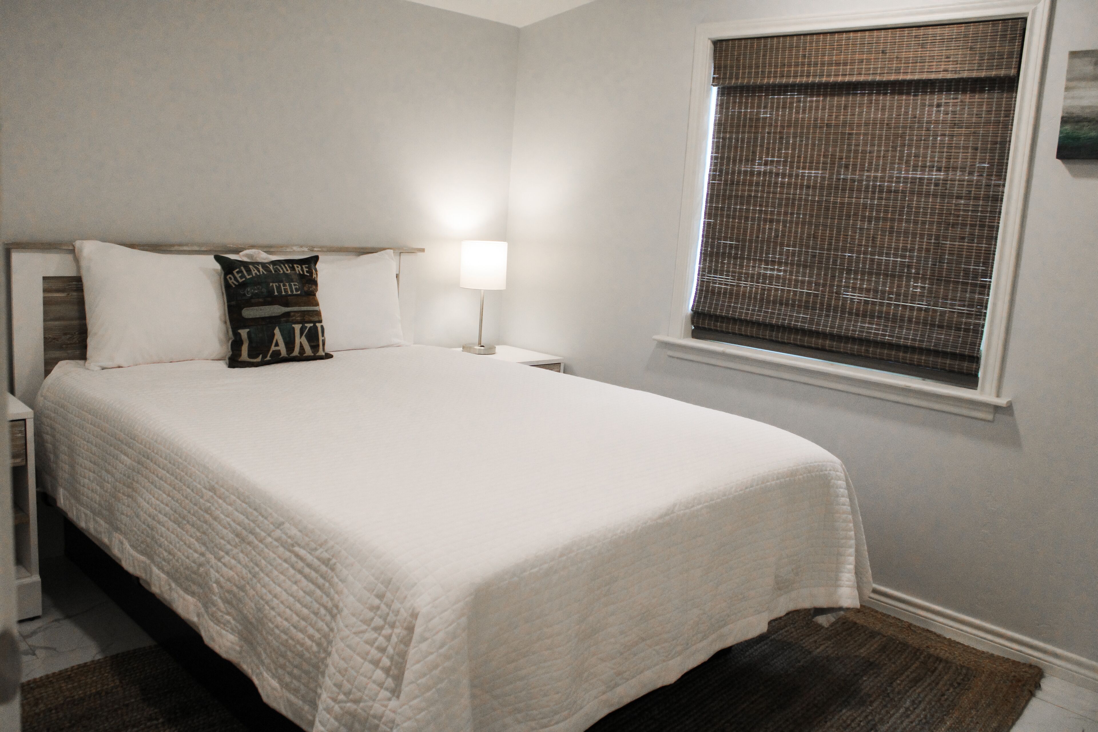 A neatly made bed with a white quilt, a pillow with text, a bedside lamp, and a window with blinds.
