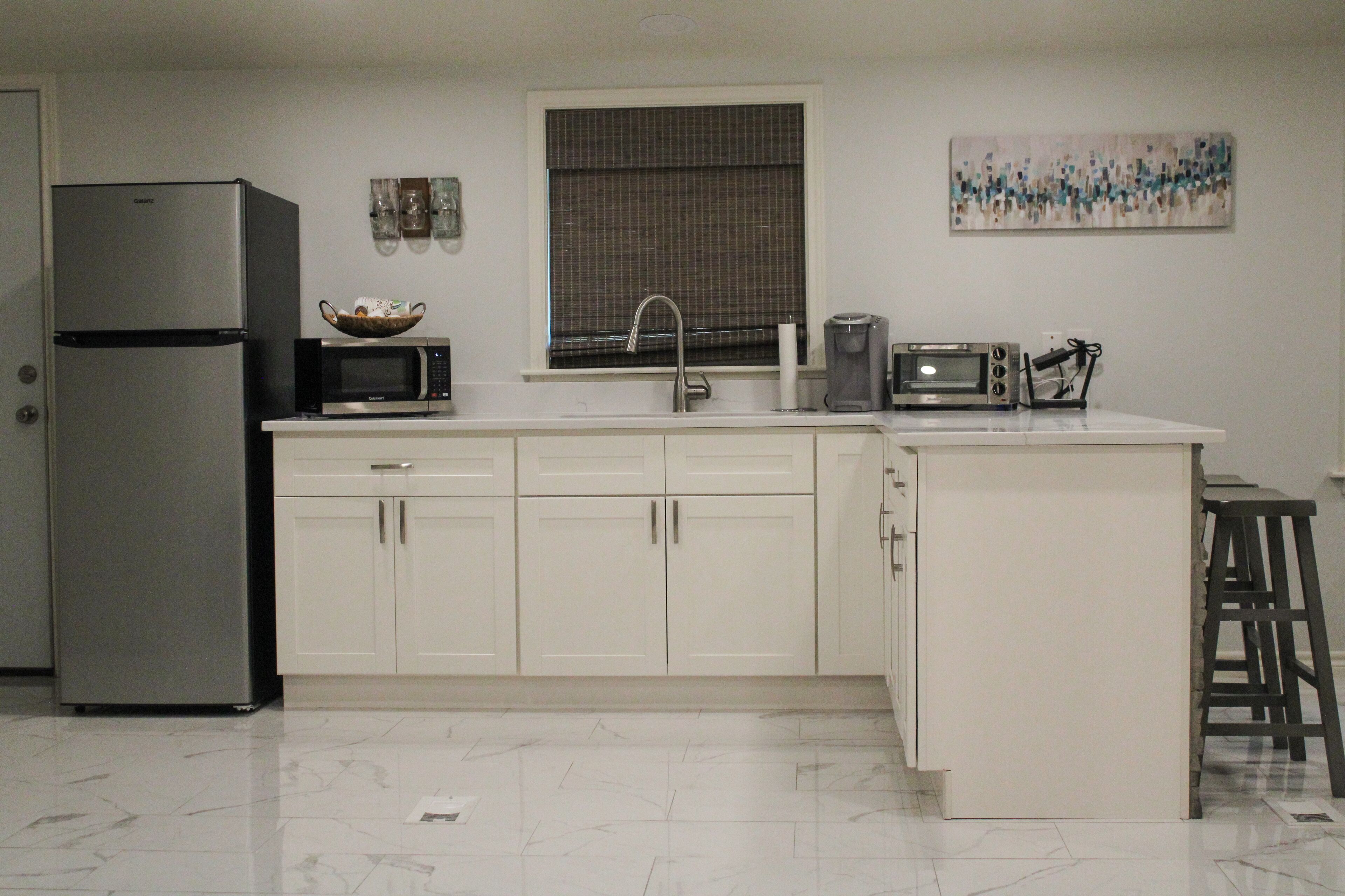 A kitchen with white cabinets, a stainless steel refrigerator, a microwave, a coffee maker, and a window with blinds.