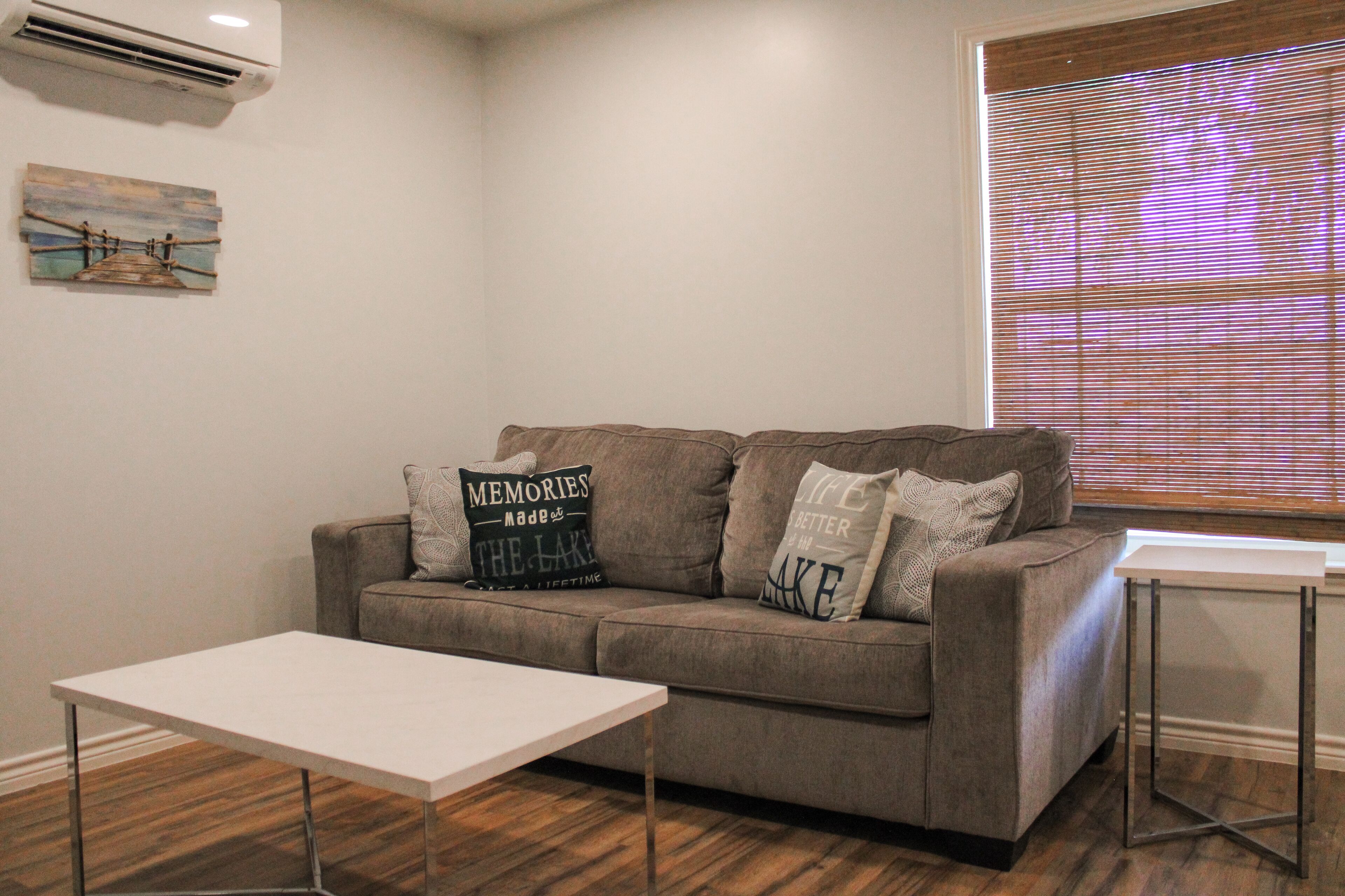 A living room with a grey sofa, a white coffee table, and a small side table. There is a painting on the wall and a window with blinds.