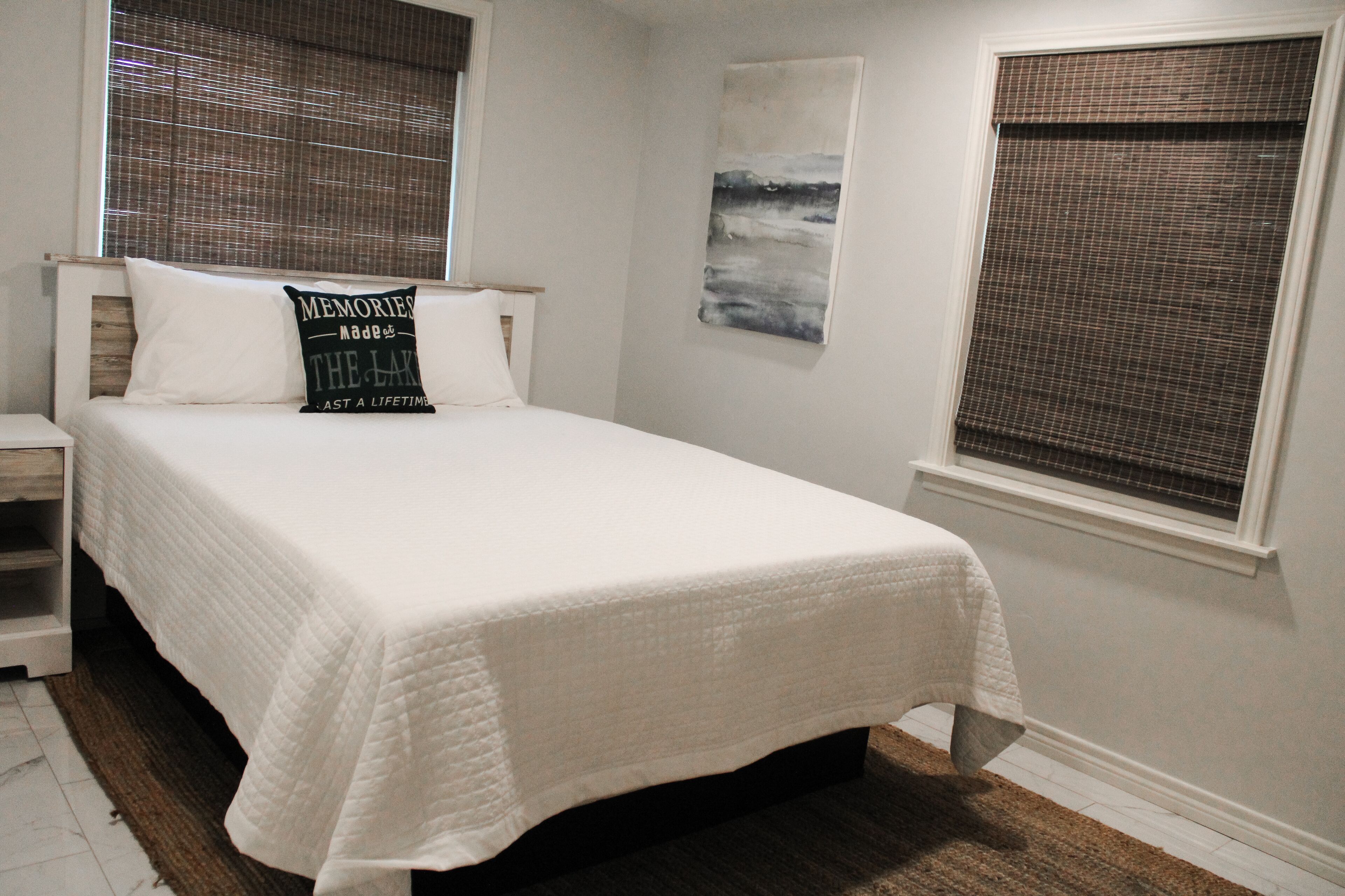 A neatly made bed with a white bedspread, a pillow with text, and a painting on the wall.