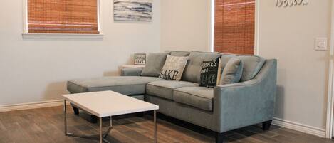 A living room with a sectional sofa, a small white table, and two windows with blinds.