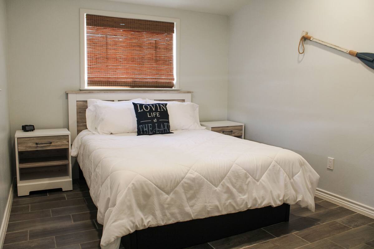 A bedroom with a bed, a nightstand, a bedside table, a window with blinds, and a wall-mounted lamp.