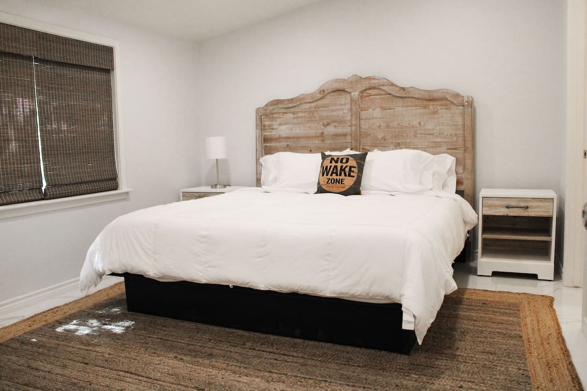 A bedroom with a wooden headboard, white bedding, a bedside table with a lamp, and a window with blinds.