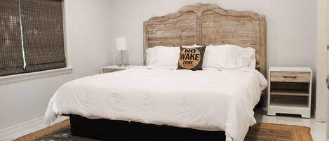 Une chambre à coucher avec une tête de lit en bois, une literie blanche, une table de chevet avec une lampe et une fenêtre avec des stores.