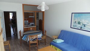 A room with a bed, a blue sofa, a wooden dining table with chairs, a kitchen area, and a white pendant light. - Hardelot Apartment Panoramic beach and sea view (Neufchâtel-Hardelot)