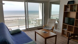 A living room with a view of the beach, a sofa, a table, a chair, and a bookshelf. - Hardelot Apartment Panoramic beach and sea view (Neufchâtel-Hardelot)