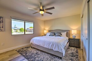 A bedroom with a bed, a ceiling fan, a nightstand, a lamp, and a window with a view of buildings and trees.