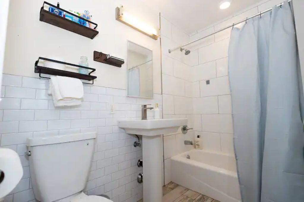 Ein Badezimmer mit Toilette, Waschbecken und Badewanne. Regale mit Pflegeprodukten und ein Spiegel über dem Waschbecken.