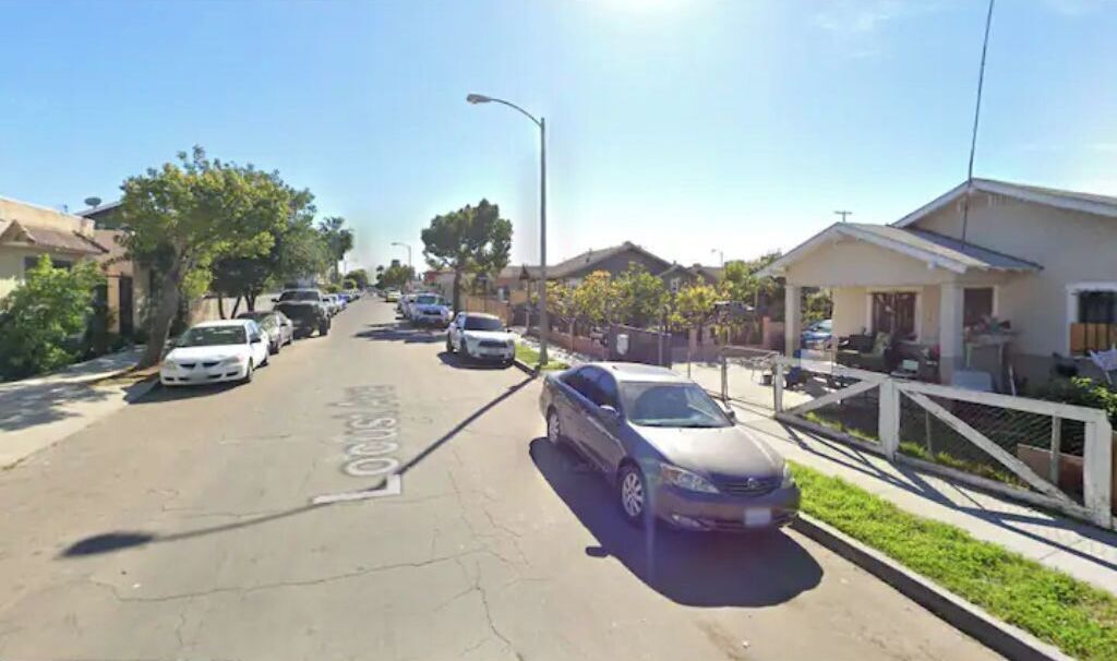 Une rue résidentielle bordée de voitures garées, de maisons et d’un ciel dégagé.