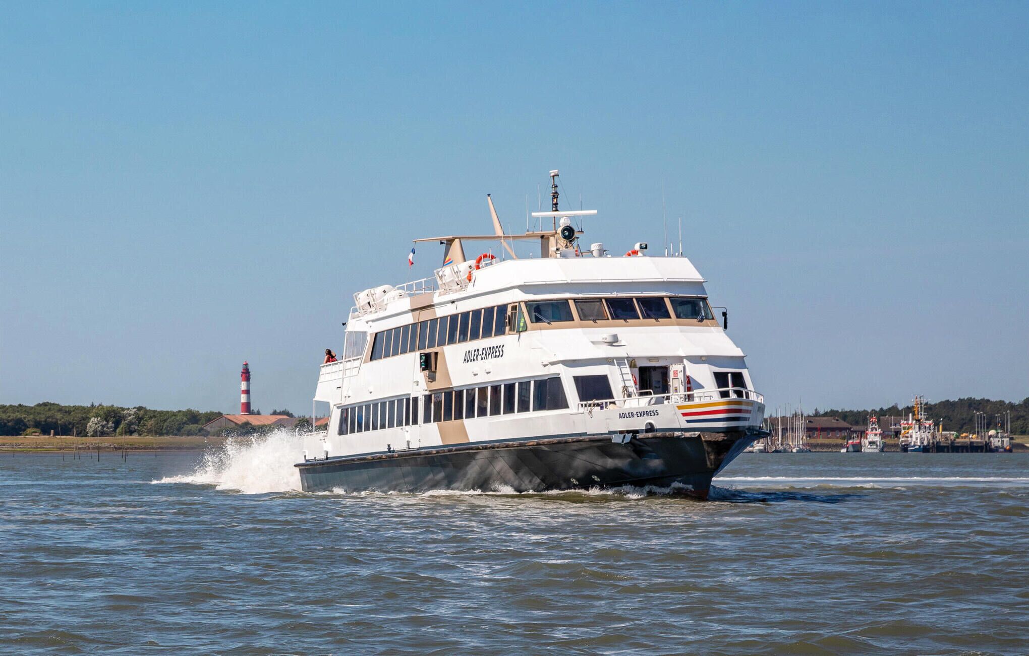 A white, multi-deck ferry with "ADLER EXPRESS" written on the side, moving through a body of water with a lighthouse in the background.