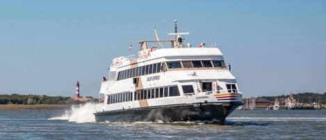 A white, multi-deck ferry with "ADLER EXPRESS" written on the side, moving through a body of water with a lighthouse in the background.