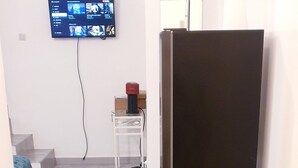 A room with a black refrigerator, a TV mounted on the wall, a small table with a vacuum cleaner, and a trash bin. - Magnificent F2 with terrace and parking, close to a park and 7 minutes from station (Le Mée-sur-Seine)