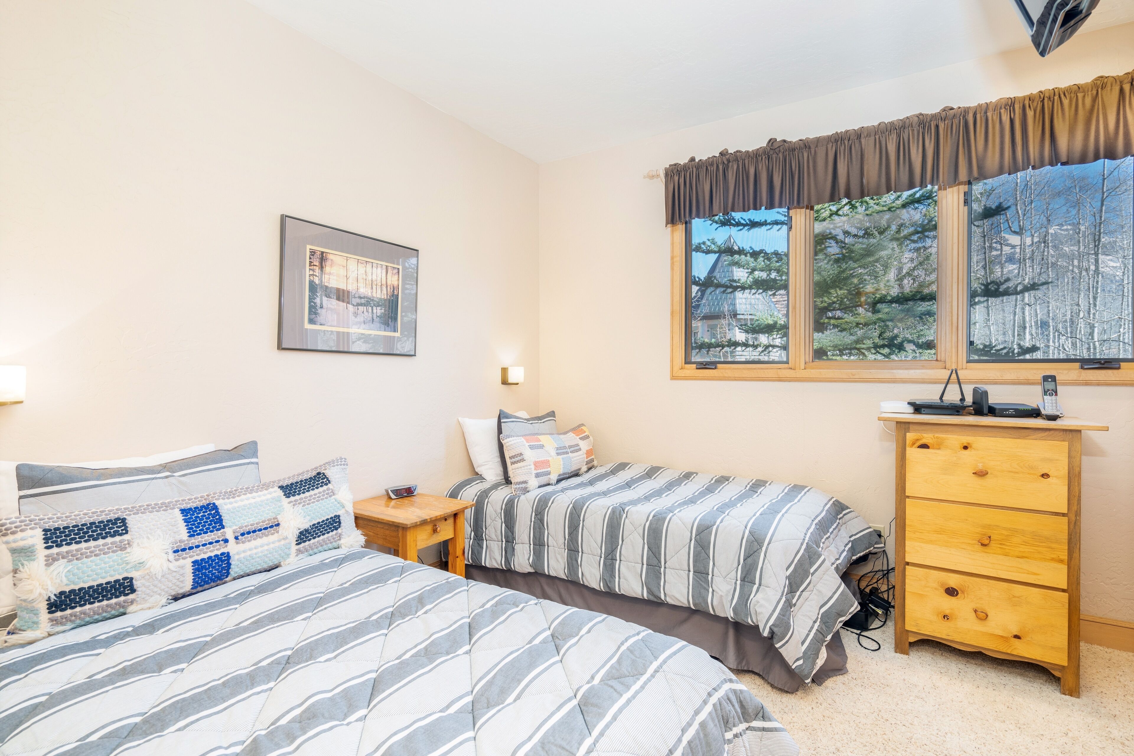 A bedroom with two beds, a dresser, a framed picture on the wall, and a window with a view of trees.