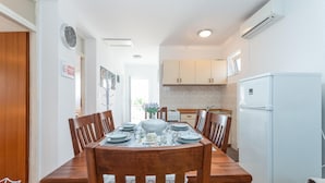 Un comedor con una mesa de madera dispuesta para cuatro personas, rodeada de sillas. Se observa un refrigerador blanco a la derecha y, al fondo, una cocina con alacenas y una estufa.