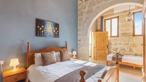 Une chambre avec un grand lit, une tête de lit en bois, une table de chevet avec une lampe, un tableau au mur et une arche en pierre menant à une autre pièce.