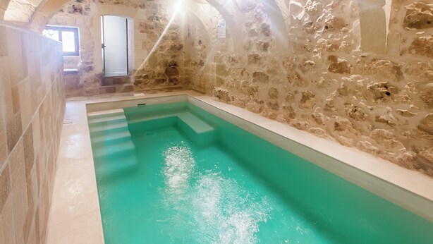 Une piscine intérieure aux murs de pierre, dotée d’une eau claire, de voûtes et d’une fenêtre laissant entrer la lumière naturelle.