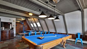 A pool table with a blue felt surface, surrounded by blue chairs, in a room with a sloped ceiling and large windows. - The Birch Ridge: Mission Room #2 - Queen Suite In Renovated Killington Lodge. Hot Tubs. 1 Bedroom Home (Killington)