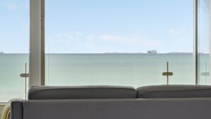 Blick aus einem Fenster mit einer Couch im Vordergrund, auf ein ruhiges Meer mit einem Schiff in der Ferne.