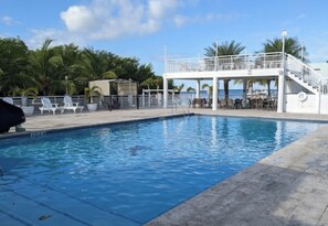 A swimming pool with a white deck, lounge chairs, and a covered area with tables and chairs.