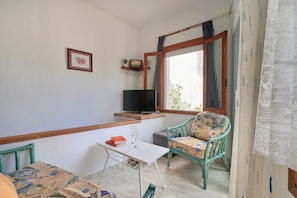 A small, cozy room with a television, a table with a book and glass, a patterned chair, and a window with curtains.