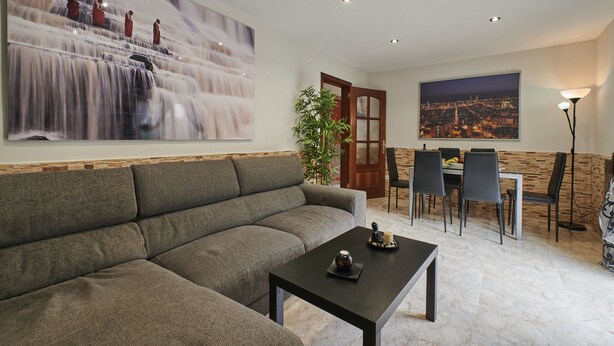 Una sala de estar con un sofá gris, una mesa de centro, un comedor con una mesa y sillas, un cuadro de una cascada y un cuadro de un paisaje urbano.