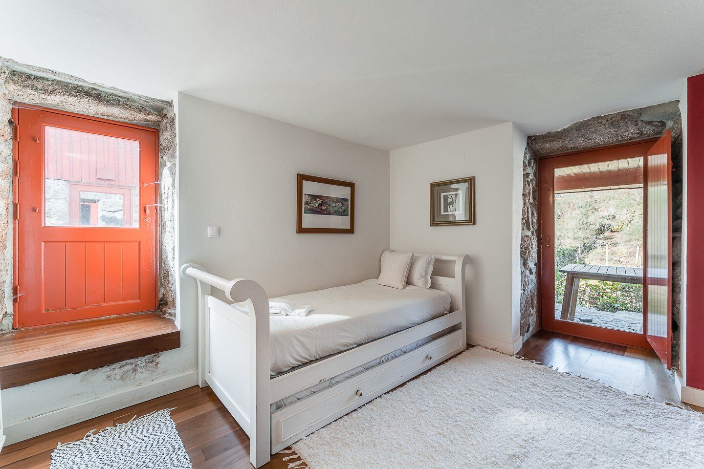 Une petite chambre avec un lit simple, une fenêtre avec un cadre rouge et une vue sur l’extérieur.