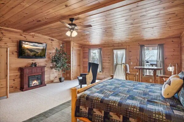 A wooden cabin bedroom with a bed, a fireplace, a ceiling fan, and a television on the wall. - Mountain views, HUGE theater room, hot tub, game room! (Sevierville)