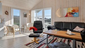 Amplia sala de estar con techo de madera, un sofá de cuero negro, un sillón blanco y una mesa de comedor con alfombra a rayas.
