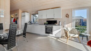 Una cocina moderna con armarios blancos, un refrigerador de acero inoxidable y un horno empotrado. Hay un comedor con una mesa y sillas de vidrio, y un ventanal que ofrece vistas al exterior.