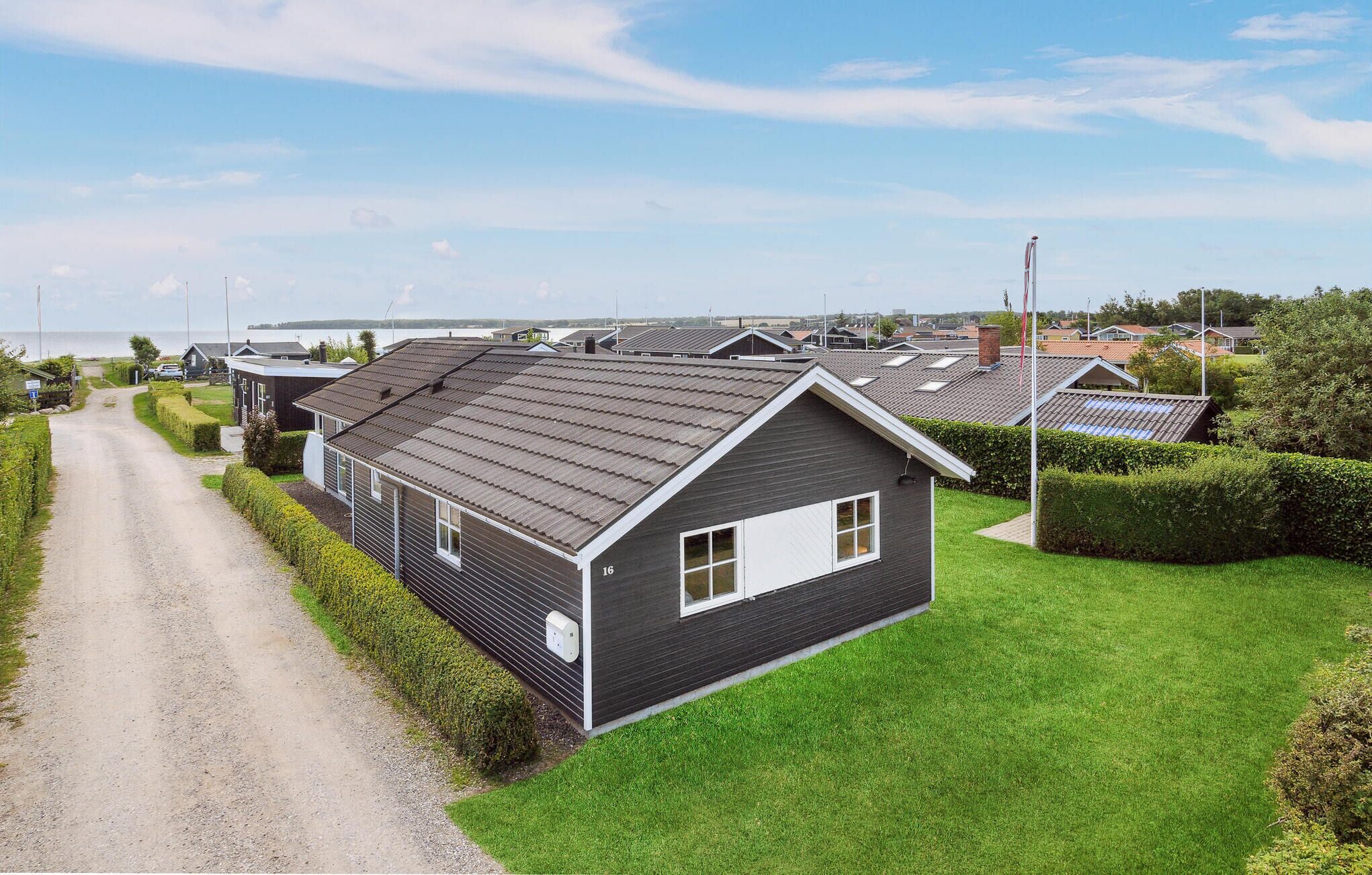 Une rangée de maisons modernes à un seul étage, avec des toits sombres et des éléments décoratifs blancs, est disposée le long d’un chemin de gravier, devant lequel se trouve une zone herbeuse.