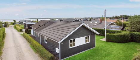 Une rangée de maisons modernes à un seul étage, avec des toits sombres et des éléments décoratifs blancs, est disposée le long d’un chemin de gravier, devant lequel se trouve une zone herbeuse.
