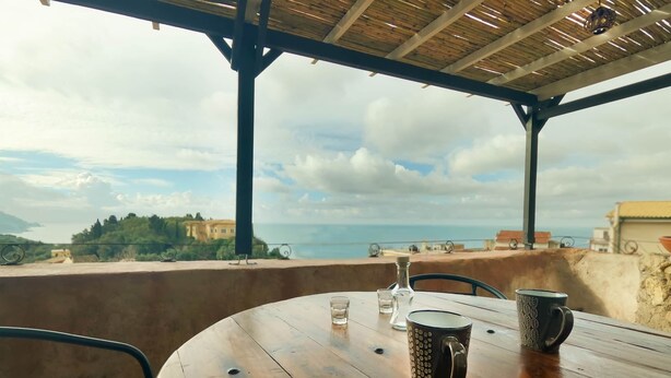 Ein Holztisch mit zwei Tassen und einem Glas, platziert auf einem Balkon mit Blick auf das Meer und Gebäude.
