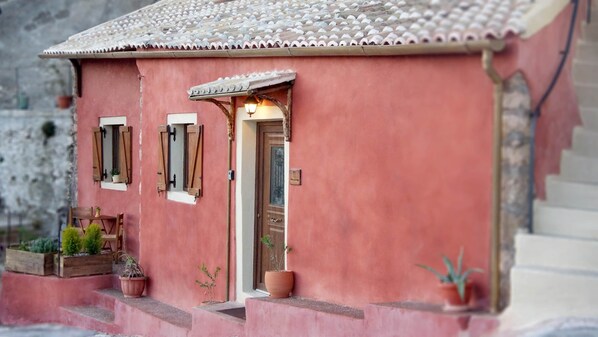 A small pink building with a tiled roof, a wooden door, and two windows with shutters. There are potted plants outside. - Papou cottage (Pelekas)