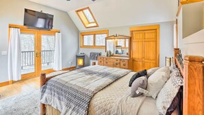 Un dormitorio con cama, ventilador de techo, televisión, chimenea, cómoda y un ventanal grande.