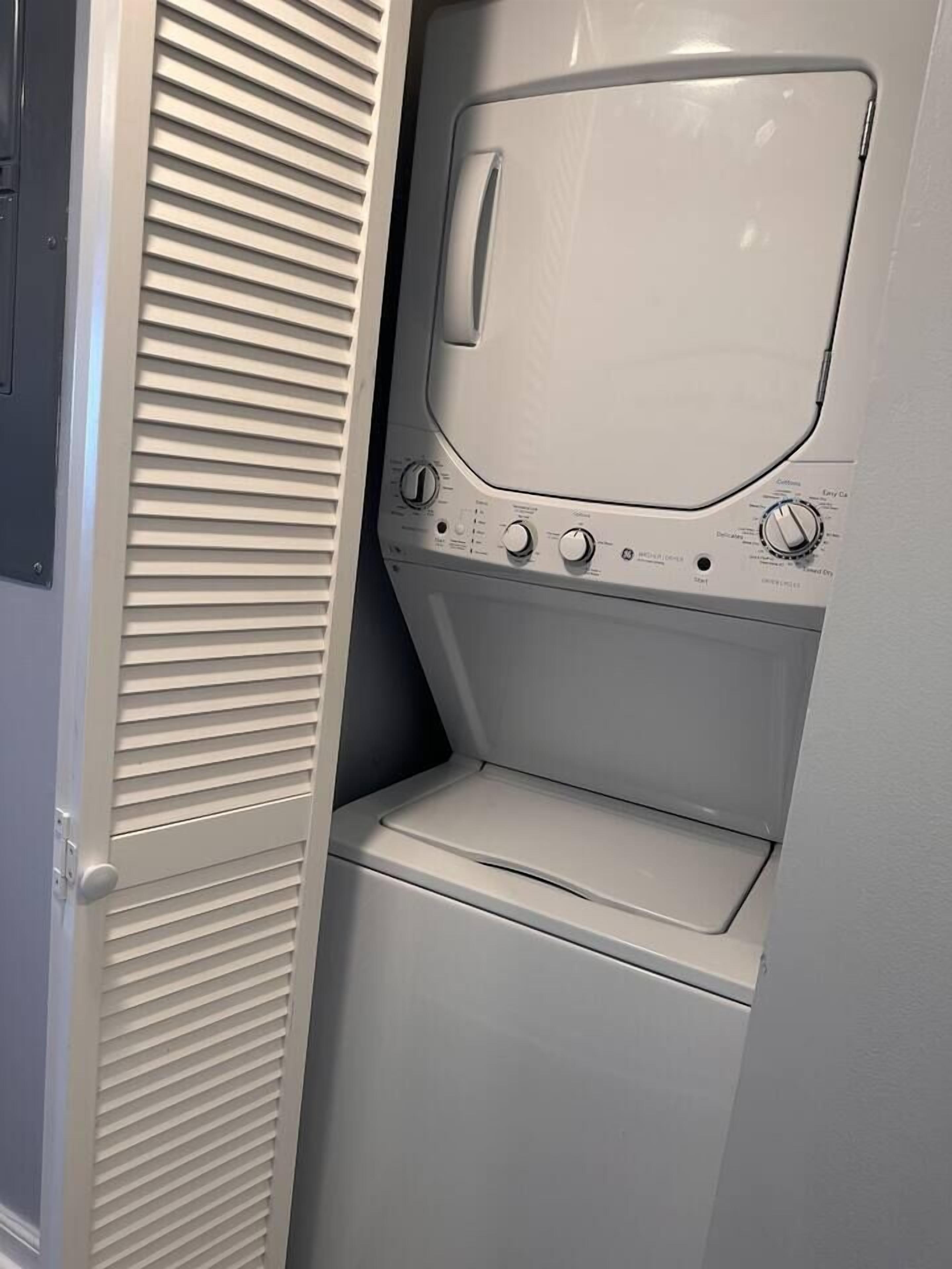 A compact, white laundry room with a built-in washer and dryer, a small shelf, and a door with horizontal louvered panels.