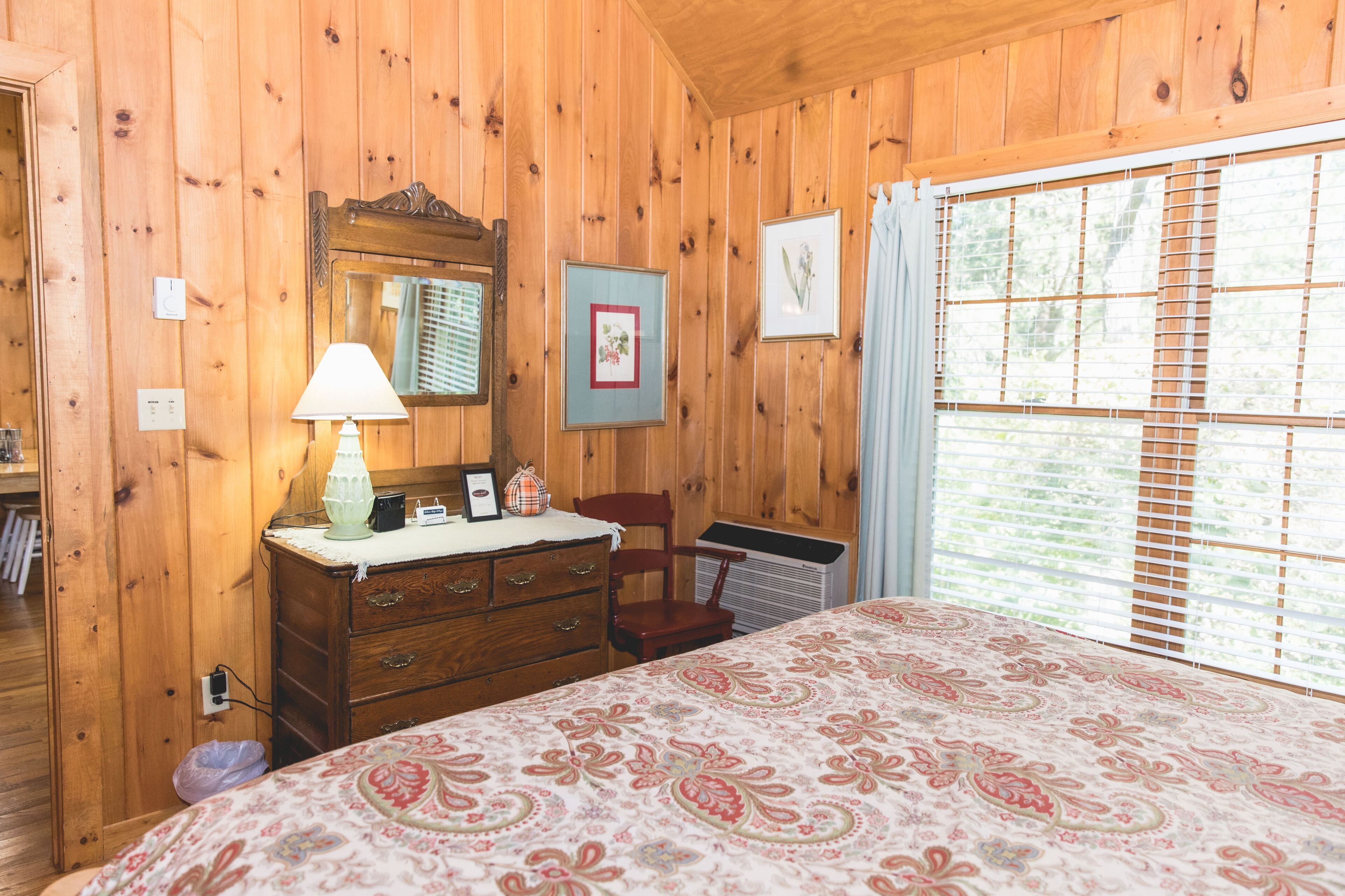 A wooden-paneled room with a bed, dresser, mirror, and window with blinds.