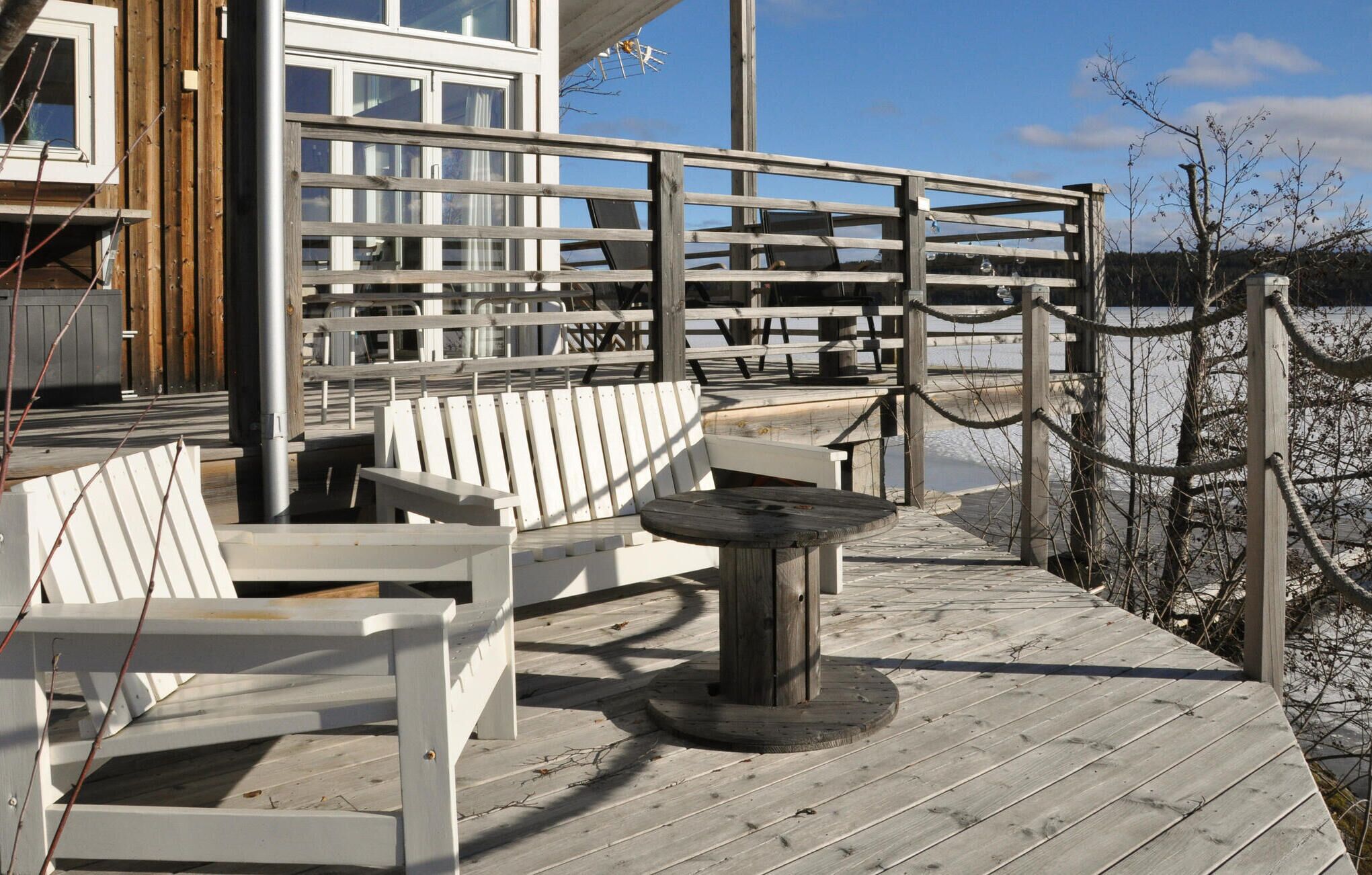A wooden deck with white Adirondack chairs and a round table, overlooking a body of water.