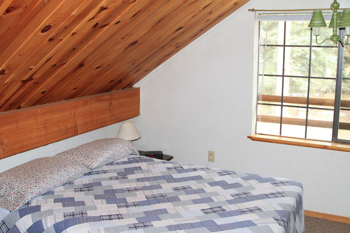 Un dormitorio con techo de madera, cama con colcha a cuadros, mesita de noche, lámpara y ventana con vistas a zonas verdes.