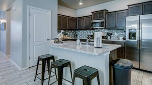 Cozinha moderna com armários de madeira escura, geladeira de aço inoxidável, micro-ondas e bancada de granito. Há três banquetas pretas e uma lixeira.